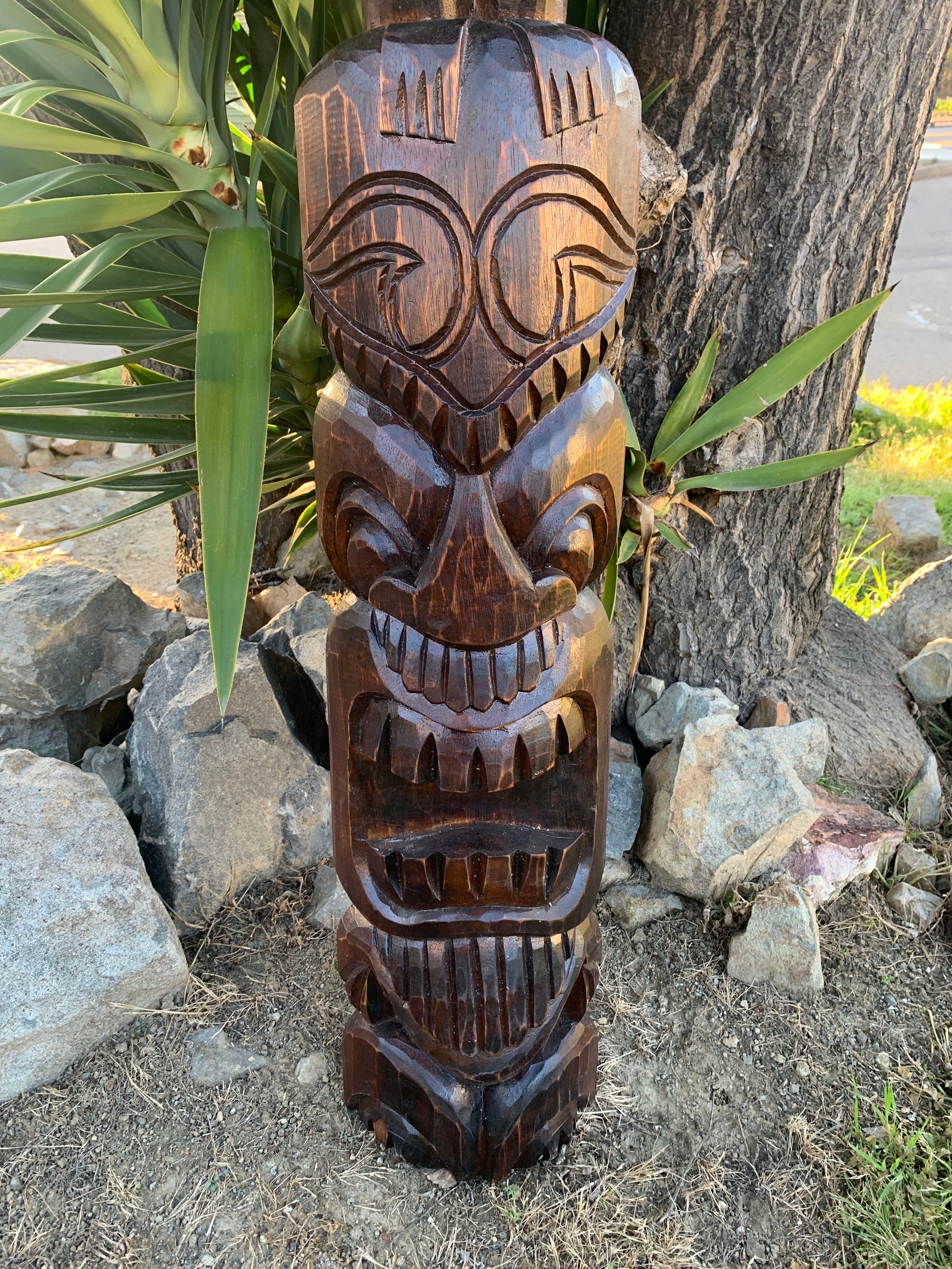 hand-carved wooden tiki statue with stacked faces beside a tree and rocks